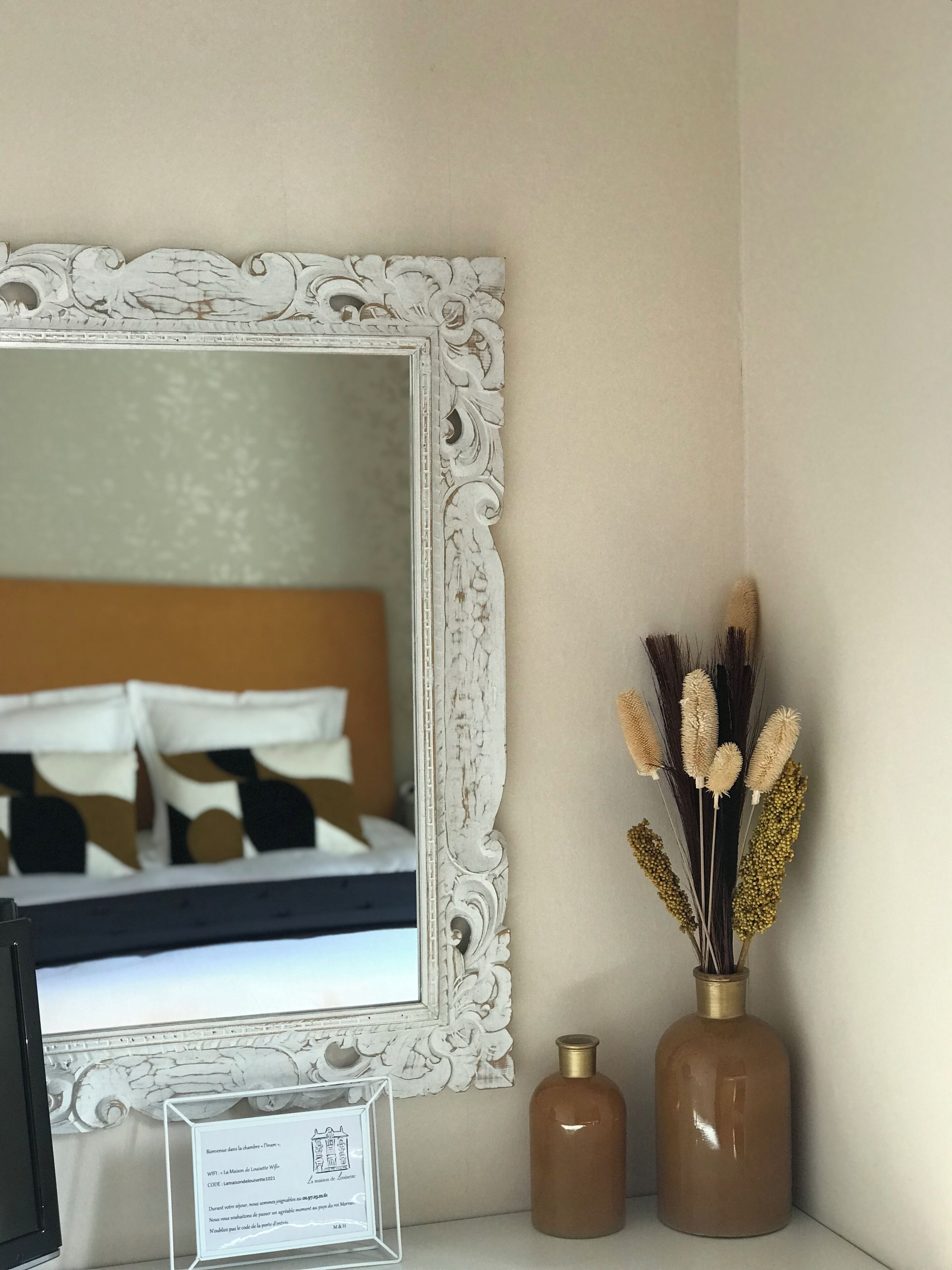 Quarto com um espelho grande e ornamentado, um certificado emoldurado e dois vasos decorativos.