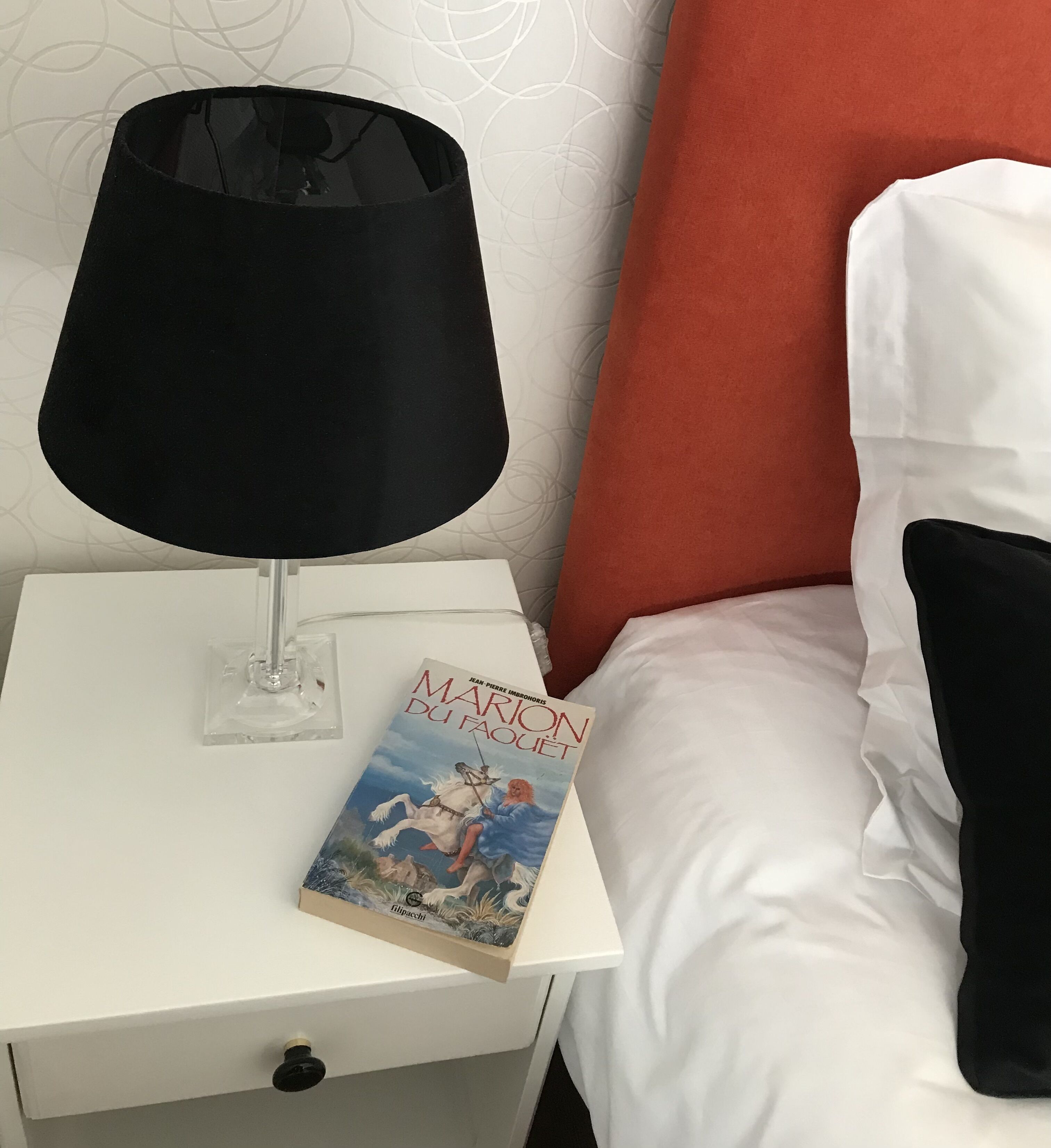 Una cama bien tendida con almohadas blancas y negras, una mesita de noche blanca con una lámpara negra, y un libro titulado 'Marion du Faouet'.