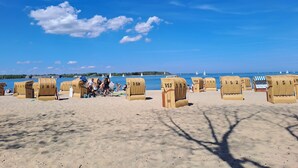 Ein Strand mit Korbstühlen für Gäste, ein klarer blauer Himmel und Segelboote auf dem Wasser.