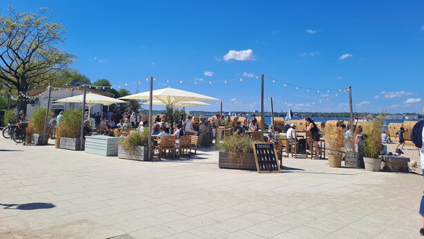 Ein Straßencafé mit Tischen und Stühlen unter Sonnenschirmen mit Blick auf ein Gewässer.