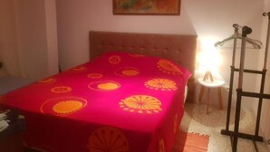 Een slaapkamer met een bed belegd met een rode deken met gele patronen, een houten hoofdbord, een klein nachtkastje met een lamp en een abstract schilderij aan de muur.