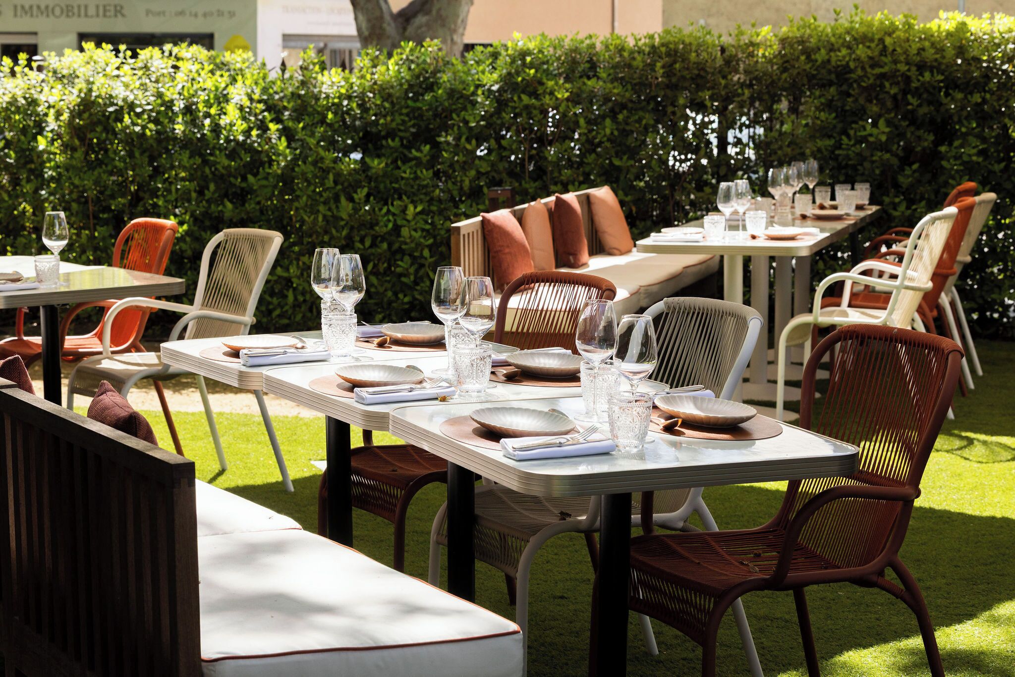An outdoor dining area with tables set for two, surrounded by chairs and a hedge.