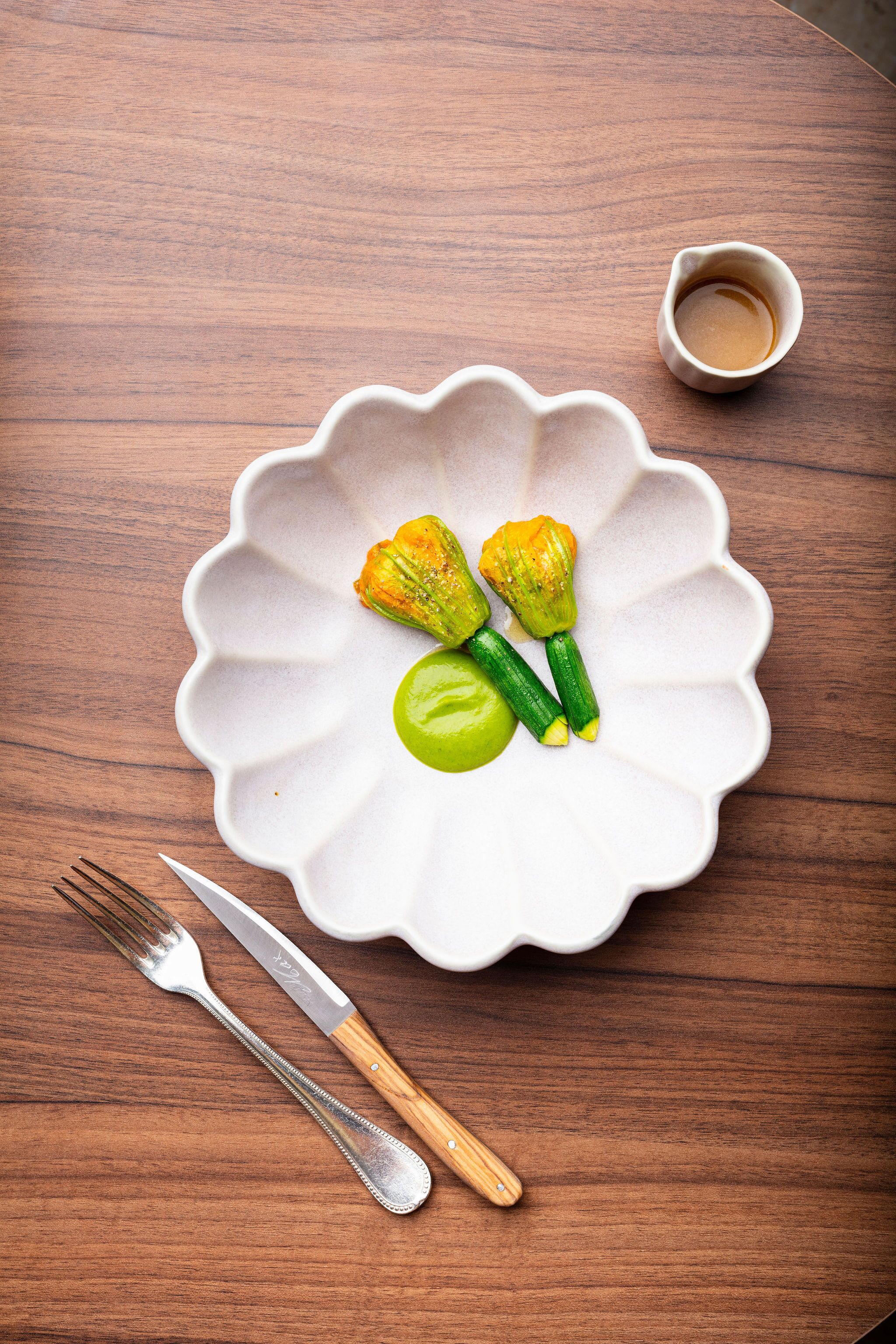 A white plate with a scalloped edge containing two green vegetables and a small bowl of sauce, placed on a wooden table.