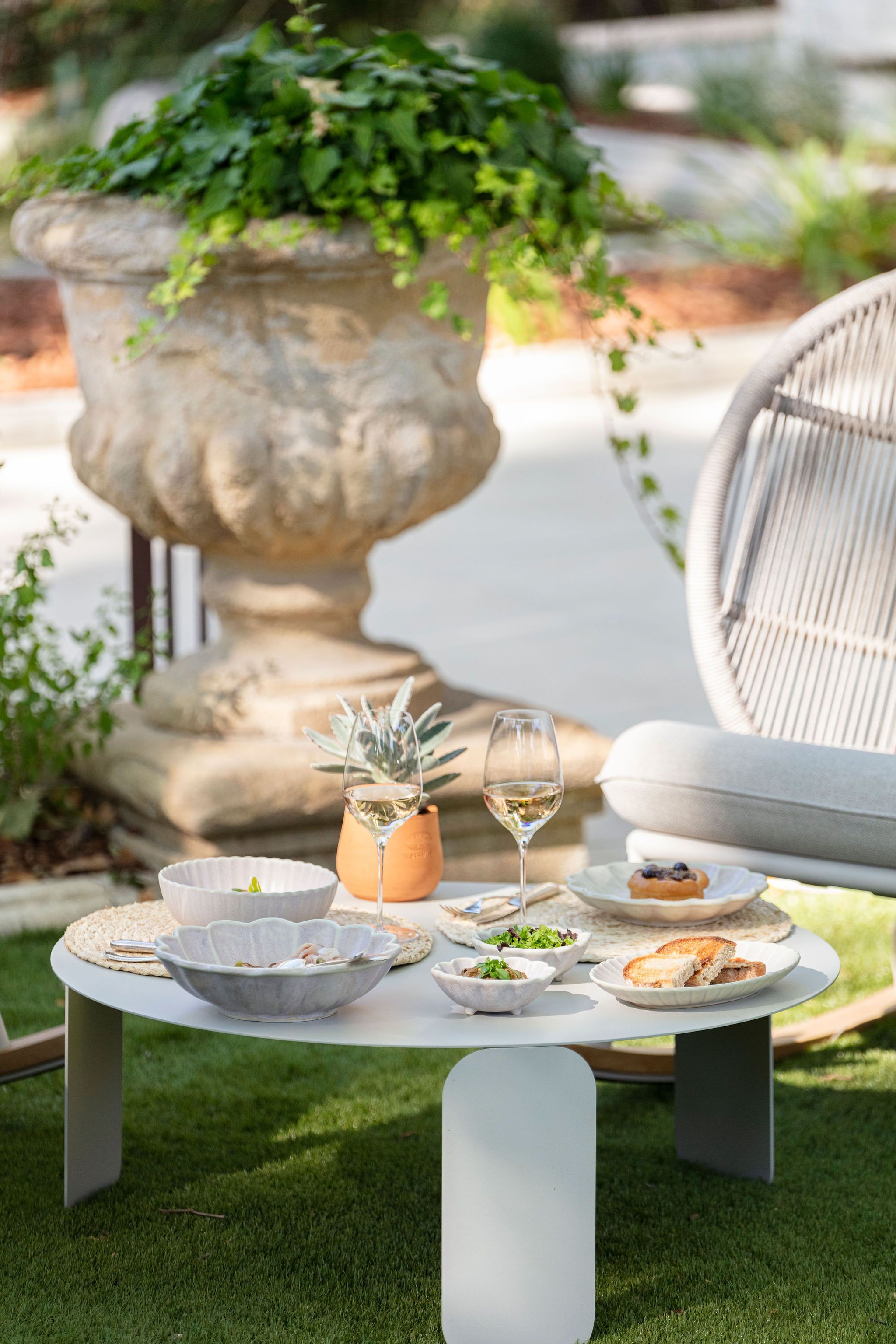 A round table with a white tablecloth, surrounded by a white chair, a potted plant, and a glass of white wine.