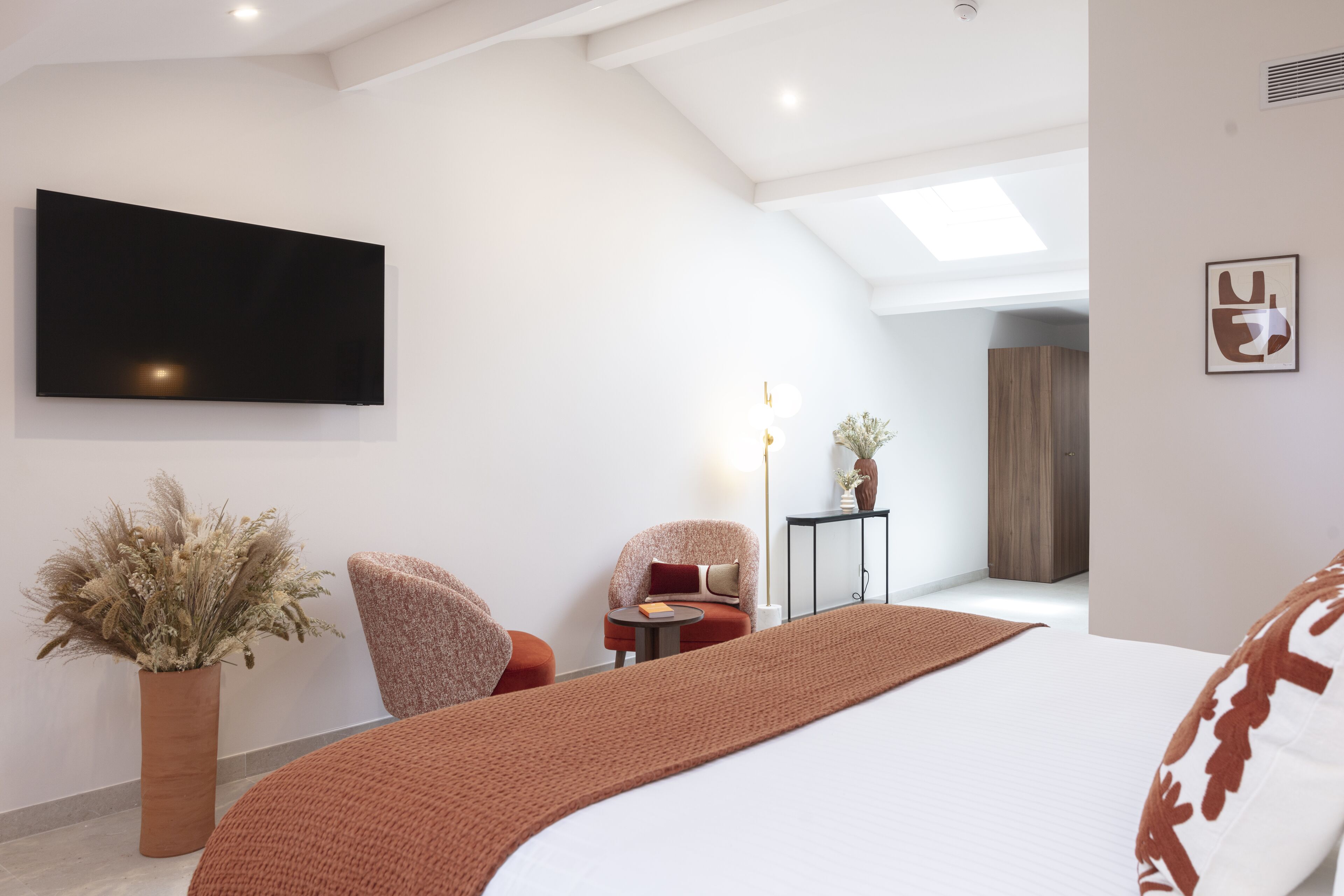 Een moderne slaapkamer met een groot bed, een flatscreen-tv aan de muur, een potplant en een zithoek met een stoel en een bijzettafeltje.