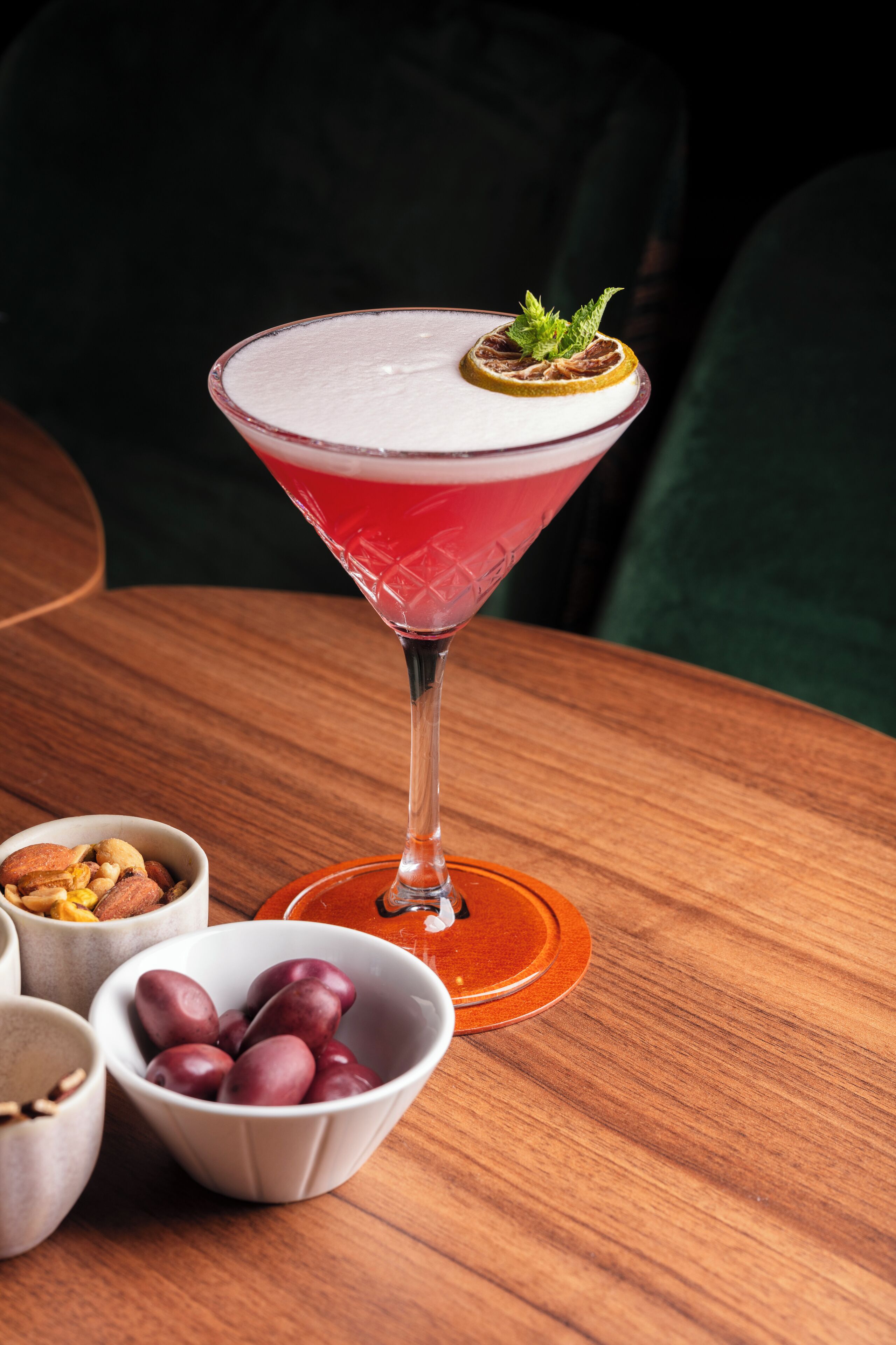 A martini glass with a red drink, garnished with a mint leaf and a slice of orange, on a wooden table.