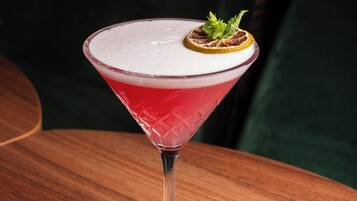 A martini glass with a red drink, garnished with a mint leaf and a slice of orange, on a wooden table.