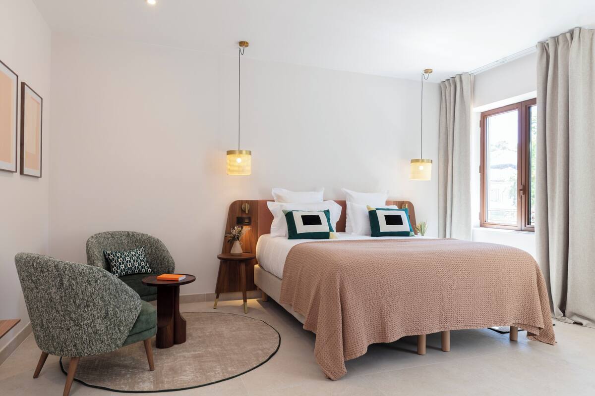 Ein modernes Hotelzimmer mit Bett, Sessel, Nachttisch und einem Fenster mit Vorhängen.
