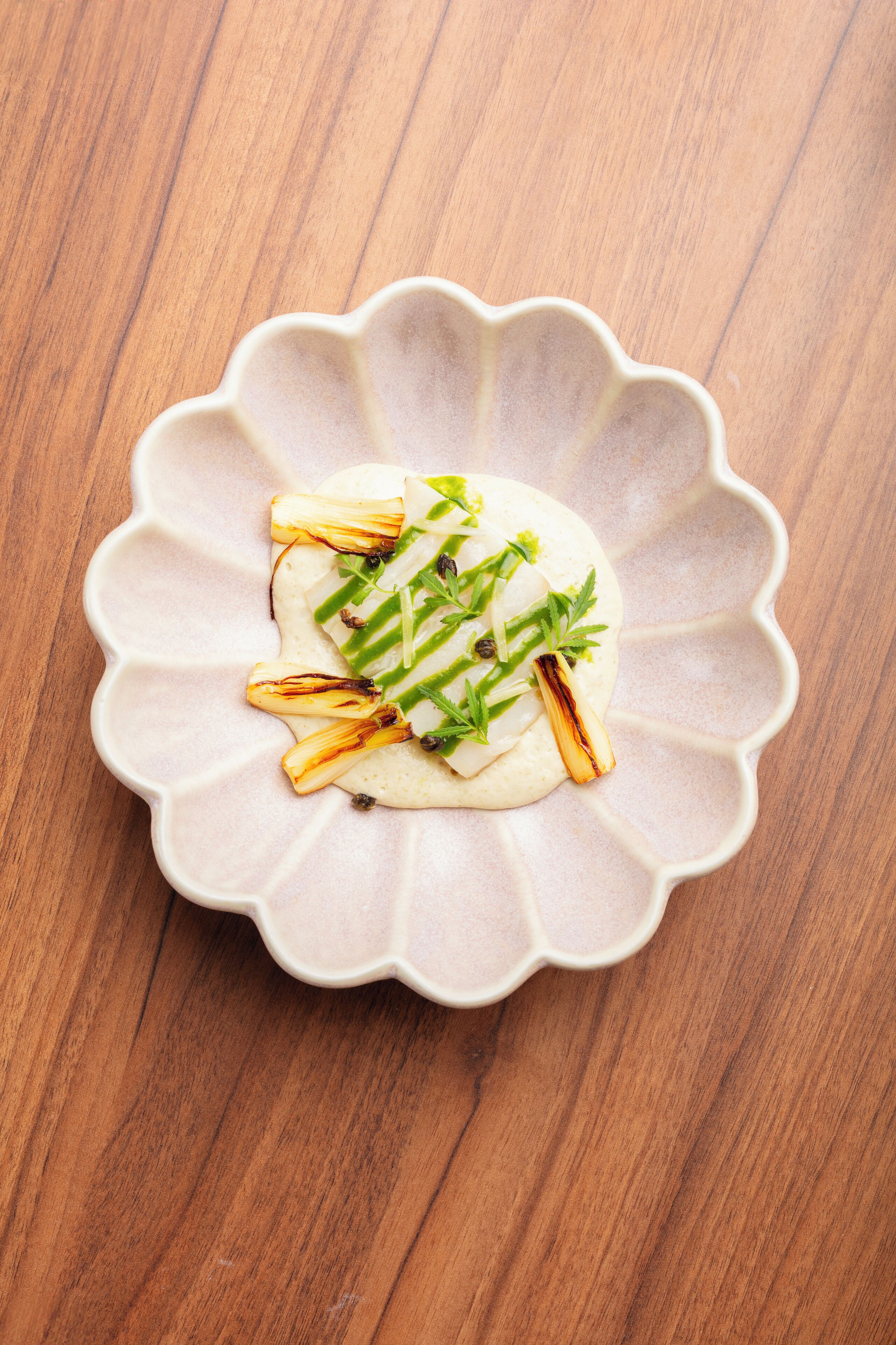 Un piatto bianco in ceramica con bordo ondulato, contenente un cibo guarnito con erbe verdi e pezzi di verdure bruciacchiate, posizionato su una superficie in legno.