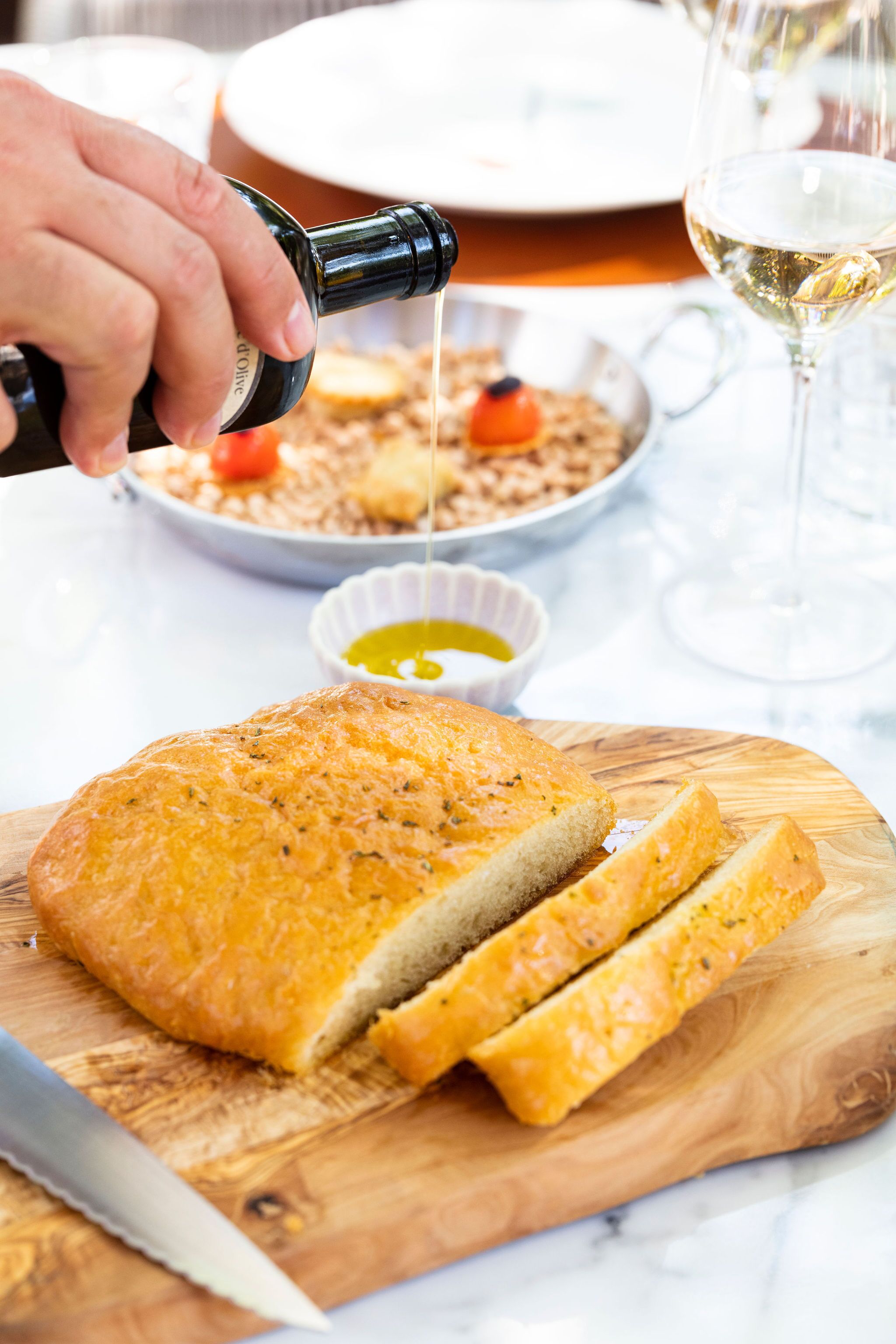 Une miche de pain sur une planche à découper en bois, avec des tranches déjà coupées, un couteau et une bouteille dont on verse le contenu dans un bol.
