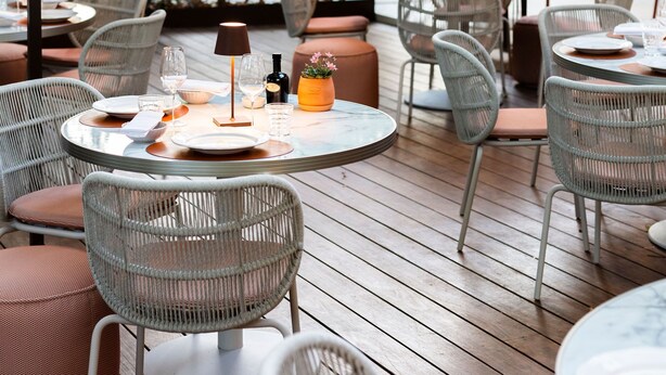 An outdoor dining area with wicker chairs and tables, surrounded by greenery and a wooden deck.