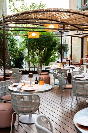 An outdoor dining area with wicker chairs and tables, surrounded by greenery and a wooden deck. - Hôtel et Restaurant La Bastide Bourrelly – Mathias Dandine (Cabries)