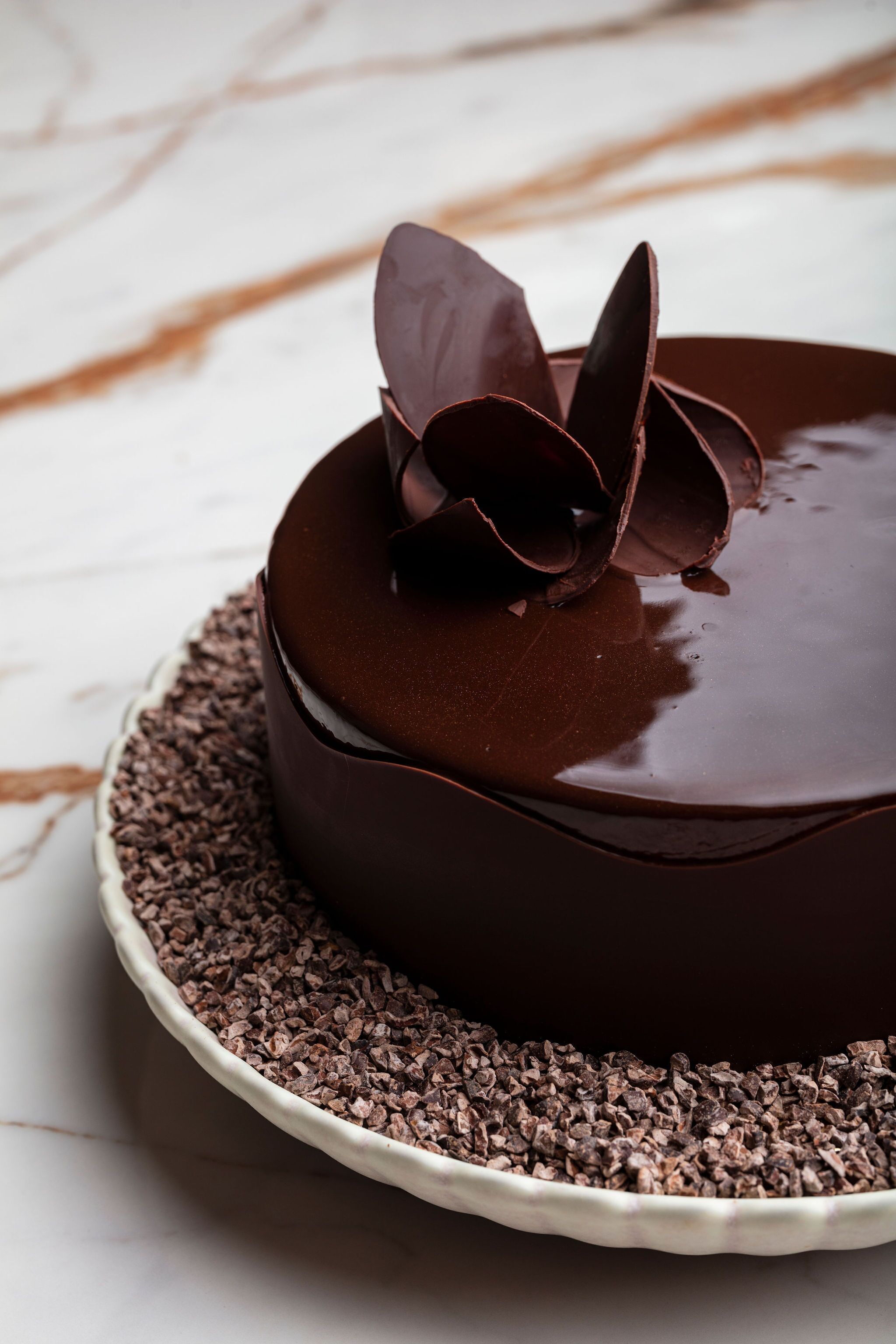 A chocolate cake with a glossy finish on a white plate, surrounded by chocolate shavings.