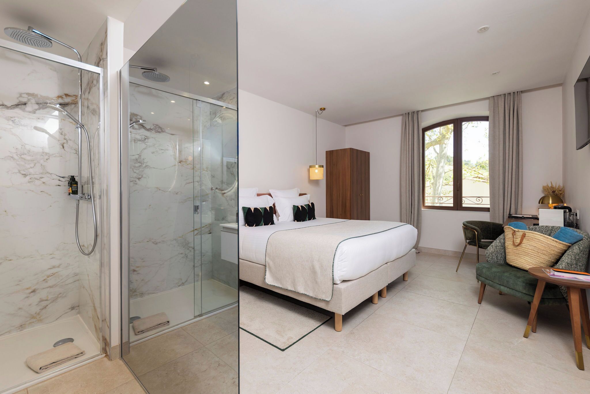 A hotel room with a glass-enclosed shower, a bed with a white duvet, a wooden wardrobe, and a view of the outdoors.