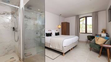 A hotel room with a glass-enclosed shower, a bed with a white duvet, a wooden wardrobe, and a view of the outdoors.