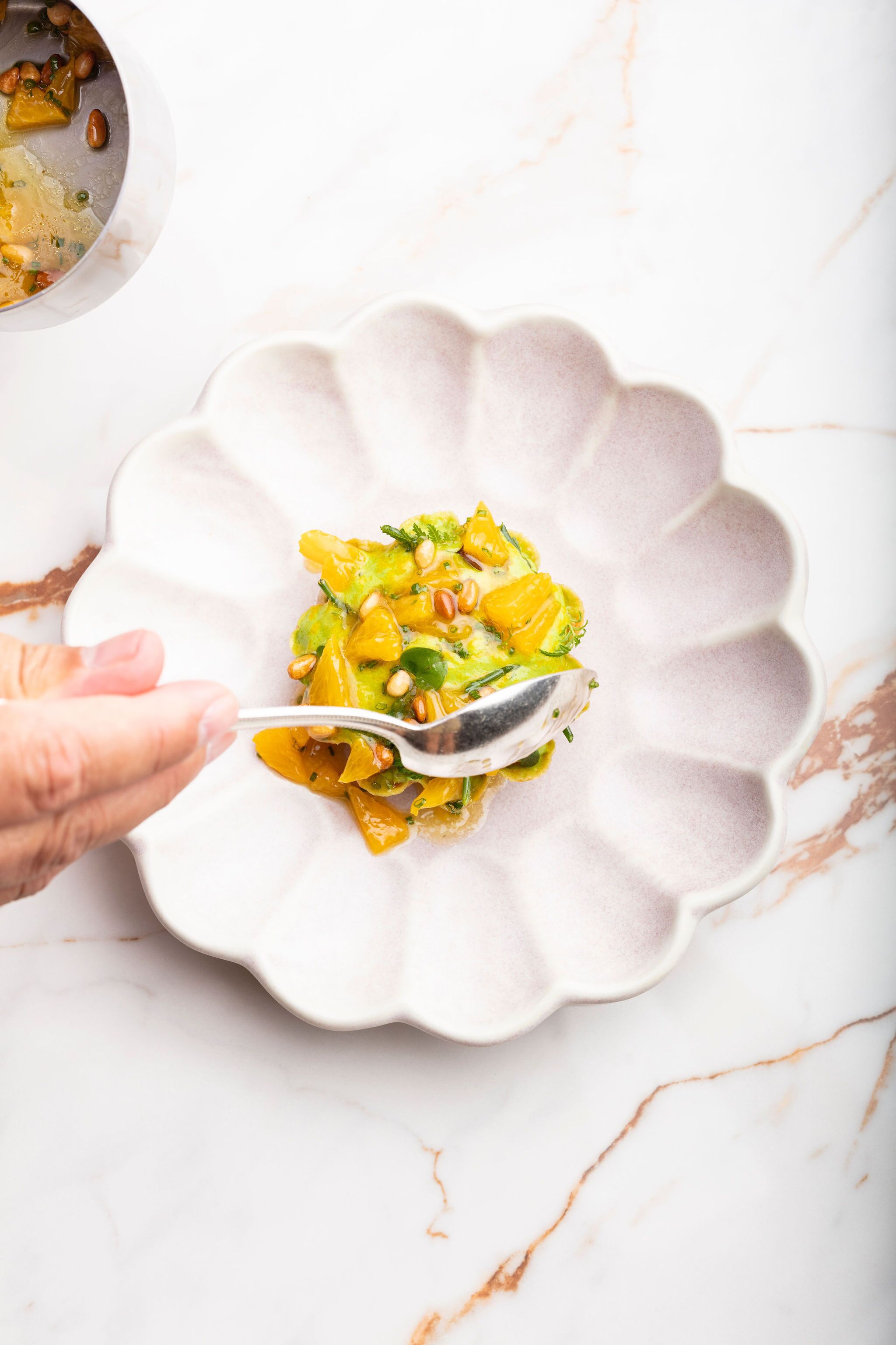 A hand holding a spoon over a scalloped white plate with a dish of food.