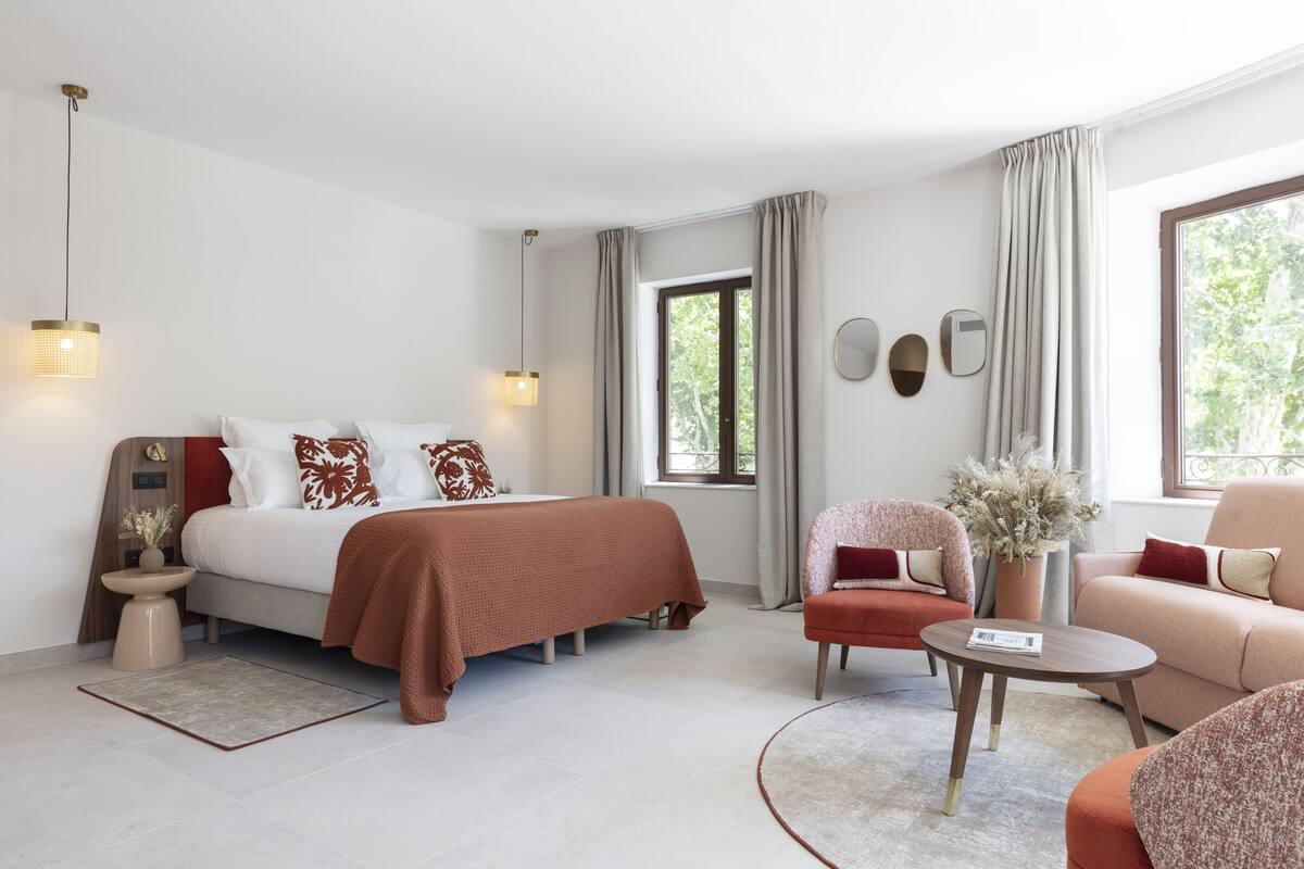 Ein modernes Schlafzimmer mit einem Bett, zwei Stühlen, einem kleinen Tisch und einem Fenster mit Vorhängen.
