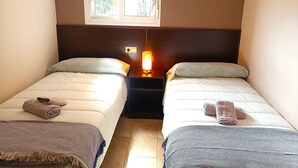 Et lille, godt oplyst værelse med to senge, et natbord med en lampe og et vindue, der giver naturligt lys.