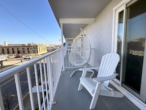 A balcony with two white Adirondack chairs and a wicker chair, overlooking a street with buildings and a clear sky. - 1BR Suite 1 Block Beach/Boards/Fun/Elevator (Ocean City)