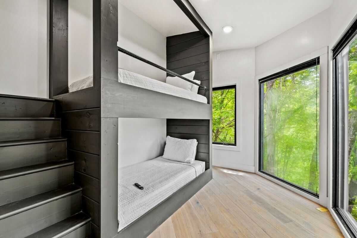 Un interno moderno con una scala, un letto con testiera e ampie finestre che danno su una vista verde.