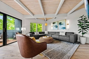 Uma sala de estar moderna com um sofá grande, uma poltrona marrom e uma mesa de centro. Há uma janela ampla com vista para o verde lá fora.