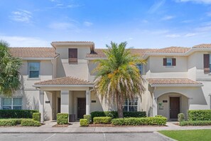 A multi-story residential building with a tiled roof, a central entrance with a covered porch, and a palm tree in the foreground.