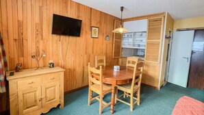 Una stanza compatta con una parete in legno, un tavolo da pranzo con quattro sedie, un piccolo armadio, un televisore a parete e un angolo cottura.