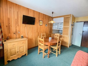 A compact room with a wooden wall, a dining table with four chairs, a small cabinet, a television mounted on the wall, and a kitchenette area. - 10m from resort center, 4th floor, balcony, tv, 37m², Tignes (Tignes)