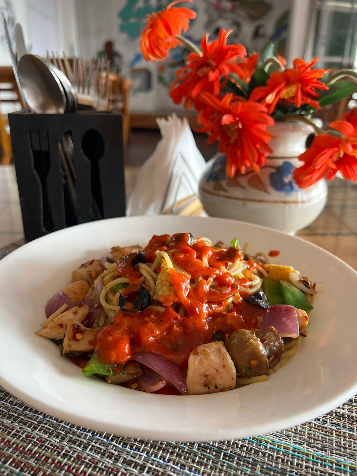 A plate of pasta with a tomato-based sauce, vegetables, and chicken, set on a table with a woven placemat.