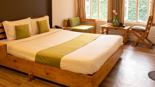 A spacious bedroom with a large wooden bed, a wooden desk, and a chair. There are windows with curtains, a wall-mounted light fixture, and a small table with a vase. - The Living Roof (Shillong)