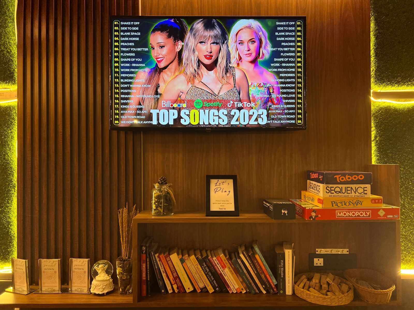 Un televisor mostrando la lista de canciones más populares de Billboard 2023 en una habitación con pared de madera, una estantería con libros y juegos de mesa.