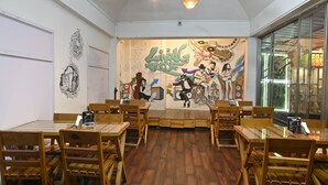 A restaurant interior with wooden tables and chairs, a mural on the back wall, and a view through a glass door. - The Living Roof (Shillong)