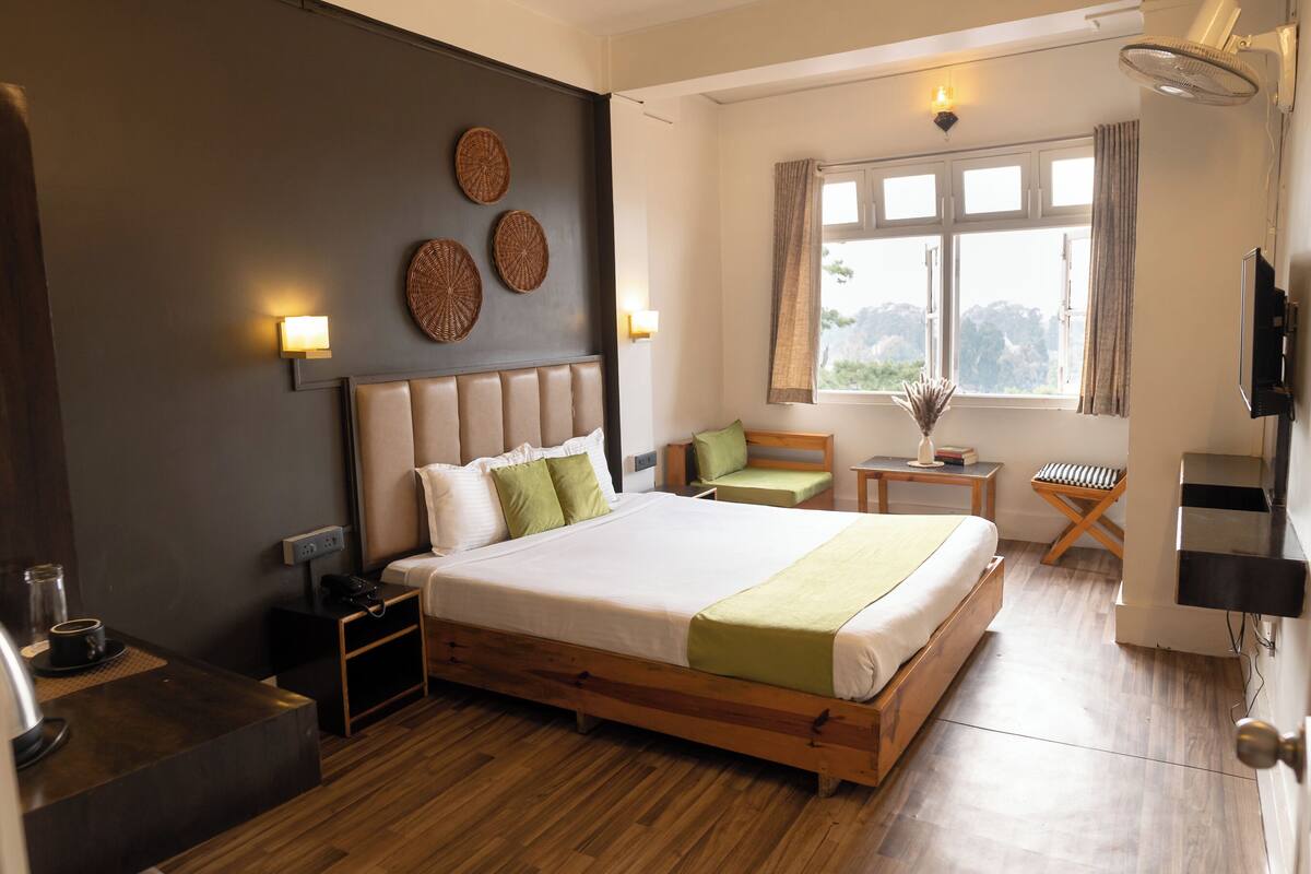 A hotel room with a large bed, bedside tables, a small seating area, a window with curtains, and a television.