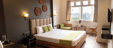 A hotel room with a large bed, bedside tables, a small seating area, a window with curtains, and a television.