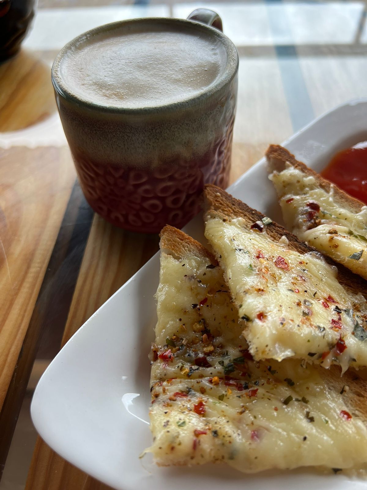 Ein Teller mit Knoblauchbrot mit geschmolzenem Käse und Kräutern, eine Kaffeetasse und eine Tomatenscheibe auf einer Holztischplatte.