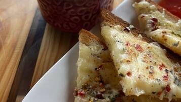 Un piatto di pane all'aglio con formaggio fuso e erbe aromatiche, una tazza di caffè e una fetta di pomodoro su un tavolo di legno.