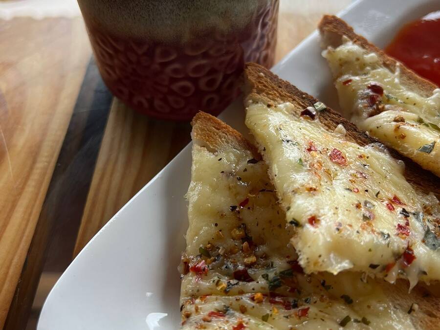 Un piatto di pane all'aglio con formaggio fuso e erbe aromatiche, una tazza di caffè e una fetta di pomodoro su un tavolo di legno.