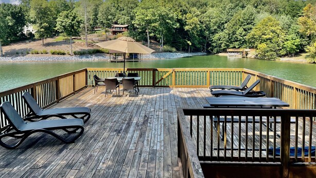 A wooden deck with lounge chairs and a table, overlooking a lake with a forested shore. - Blue Heron Point, Lakefront Luxury - Views, Party Deck, Docks, Hot Tub & FirePit (Moneta)