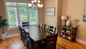 A dining room with a wooden table and chairs, a chandelier, a window with blinds, a wall-mounted sign, and a small shelf with decorative items. - Blue Heron Point, Lakefront Luxury - Views, Party Deck, Docks, Hot Tub & FirePit (Moneta)