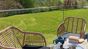 Un balcone con sedie in vimini, un tavolino in vetro e vista su un prato verde e alberi.
