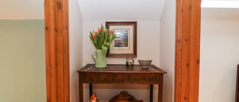 A corner room with a wooden table, a framed picture, and a vase of flowers.