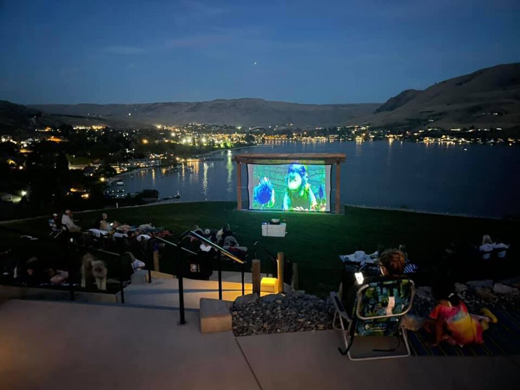 An outdoor movie screen with a group of people seated on a grassy area facing a lake, with city lights in the background.