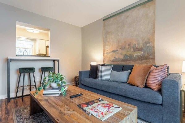 Un salon avec un canapé gris, deux tabourets de bar noirs, une table basse en bois sur laquelle sont posés des magazines, et un grand tableau abstrait accroché au mur.