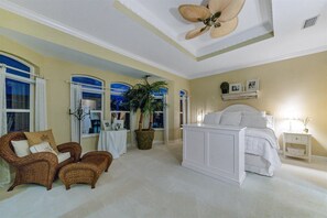 A bedroom with a large bed, a chair, a table with a plant, and a ceiling fan.