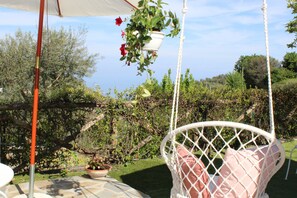 A white wicker chair with a cushion, a hanging plant with red flowers, and a table with a white umbrella. - Minicasa con ampio spazio verde attrezzato (massa lubrense)