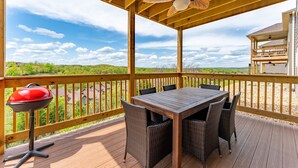 Een overdekt terras buiten met een houten tafel en stoelen, een barbecue en uitzicht op groene heuvels.
