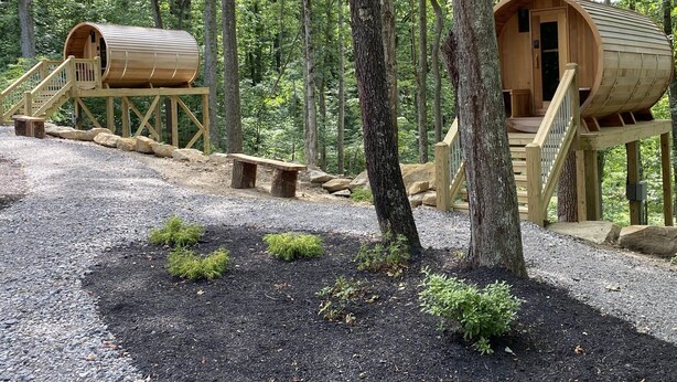 Two wooden cylindrical cabins with wooden staircases, set in a forest with a gravel path and small shrubs. - Legends Lane B - Hosted by The Chalets (Logan)