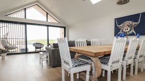 A spacious dining area with a large wooden table surrounded by white chairs, a high ceiling with skylights, and a view of the outdoors. - 5 Longshore (Newquay)