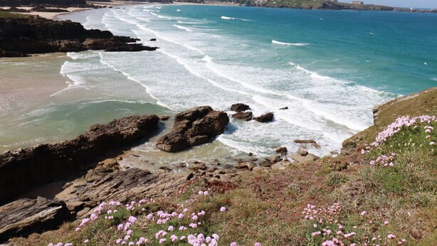A coastal landscape with rocky terrain, clear blue sea, and distant buildings. - 5 Longshore (Newquay)