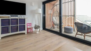 A modern living room with a large TV, a bookshelf, a floor lamp, and a view of the outdoors. - 5 Longshore (Newquay)